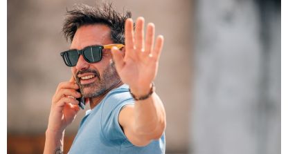 A man wearing sunglasses and a light blue T-shirt talks on a cellphone while holding up his hand toward the camera in a “stop” gesture outdoors.