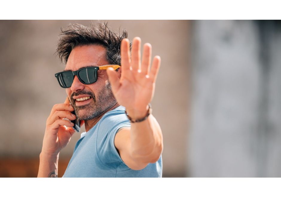 A man wearing sunglasses and a light blue T-shirt talks on a cellphone while holding up his hand toward the camera in a “stop” gesture outdoors.