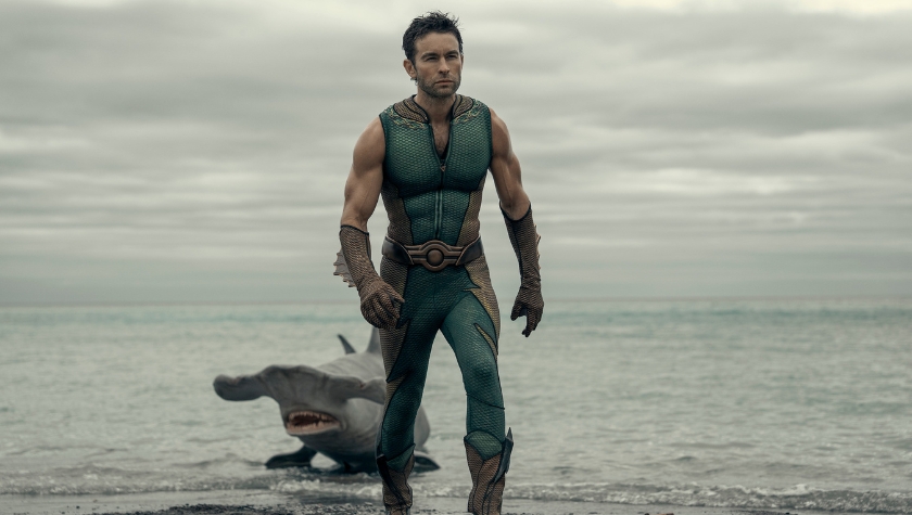 The Deep standing on a beach in his superhero costume with a shark behind him