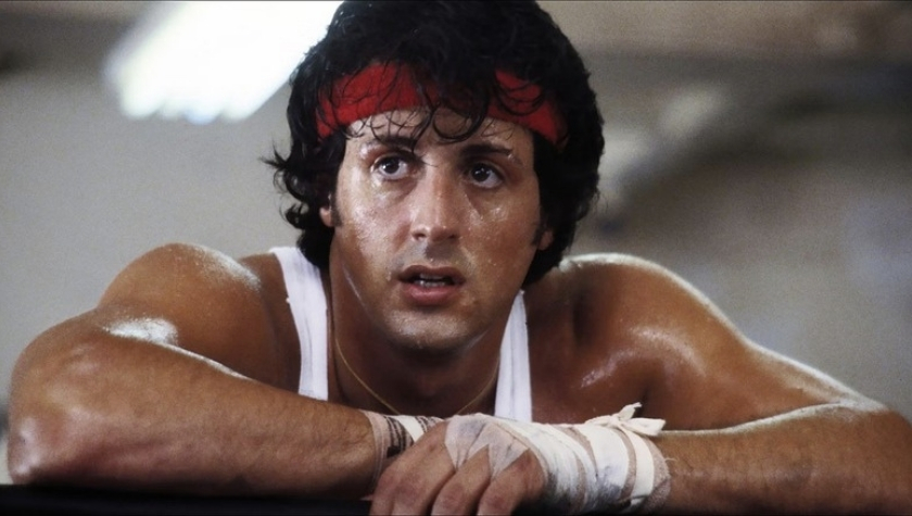 Sylvester Stallone as Rocky Balboa, leaning on the ring ropes in a boxing gym wearing a red headband and hand wraps