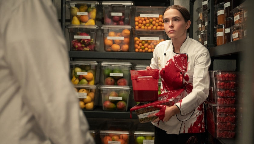 Zoey Deutch as Jill in a white chef's coat covered in red sauce, standing in a walk-in pantry storeroom