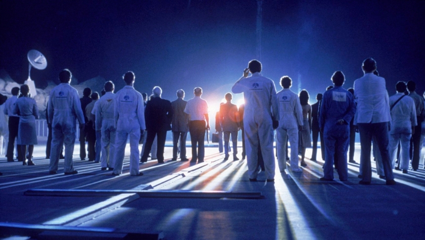 A crowd of people in white jumpsuits gathered on a tarmac, silhouetted against a blinding light in Close Encounters of the Third Kind